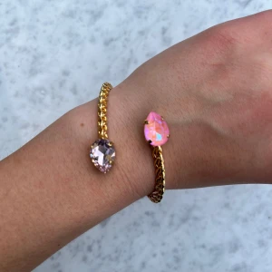 Guldigt armband med rosa stenar - Snyggt guldigt armband med tvinnad design och två droppformade stenar i rosa och ljusrosa. Stenarna har en glittrig finish som fångar ljuset och ger en lyxig känsla. Perfekt accessoar för att lyfta din outfit med lite extra färg och bling. 