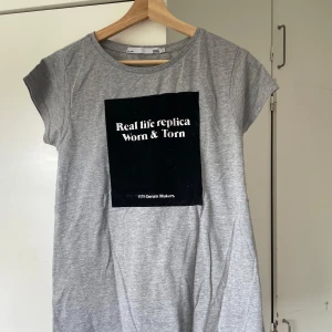 Grå t-shirt - Grå t-shirt med svart tryck framtill. Klassisk rund hals och korta ärmar. Skön och enkel modell i mjuk bomull, perfekt till jeans eller shorts.