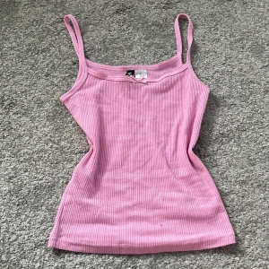 Rosa ribbad linnetopp från H&M - Supersöt ljusrosa ribbad linnetopp från H&M med smala axelband och en liten rosett framtill. Toppen är figurnära och har en mjuk, stretchig känsla som passar perfekt till jeans eller kjol. Enkel men ändå gullig detalj med rosetten vid halsringningen.
