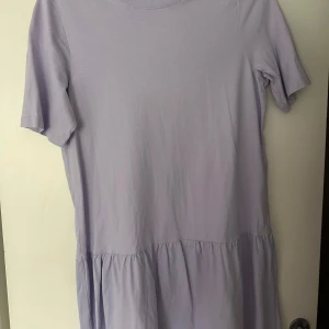 Ljuslila kort klänning i bomull - Supersöt ljuslila klänning med kort ärm och avslappnad passform. Klänningen har en rund halsringning och en volang nedtill som ger en extra touch. Perfekt för varma dagar och enkel att styla med sneakers eller sandaler.