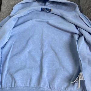 Ljusblå hoodie från Polo Ralph Lauren - Säljer en ljusblå hoodie från Polo Ralph Lauren med dragkedja, huva med snörning och klassisk logga på bröstet. Tröjan har kängurufickor och ribbade muddar. Perfekt för chill dagar och enkel att matcha med jeans eller joggers.Helt ny 