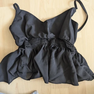 Svart smockad linnetopp från SHEIN - Svart linnetopp från SHEIN med smala axelband och smockad midja. Toppen har en luftig, volangig nederdel och är tillverkad i ett lätt, syntetiskt tyg. Perfekt för dig som gillar en enkel men ändå trendig look. Passar även XS