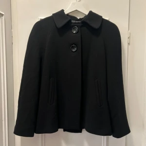 Svart kappa med stora knappar - Stilren svart kappa med klassisk krage och två stora knappar framtill. Jackan har en rak och något utsvängd passform med diskreta fickor på sidorna. Perfekt för dig som gillar en tidlös och clean look.