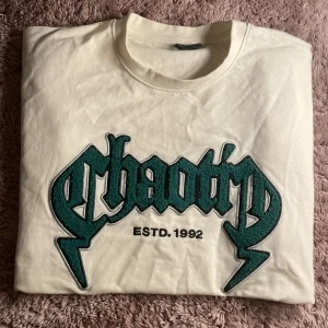 Vit oversized t-shirt från FSBN - Vit oversized t-shirt från FSBN med stort grönt broderat 'chaotic' tryck och texten ESTD. 1992 på bröstet. Klassisk rund hals och korta ärmar. Tillverkad i mjuk bomull, perfekt för en chill och avslappnad stil.