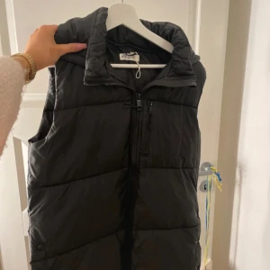 Svart dunväst med huva - Säljer en svart dunväst från H&M med huva och dragkedja framtill. Västen har en bröstficka med dragkedja och två sidofickor med knappar. Perfekt att slänga över en hoodie för extra värme under kyliga dagar. Storlek 170 och är som en S/M