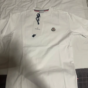 Vit Moncler t-shirt med logga - Vit t-shirt från Moncler med klassisk rund hals och broderad logga på bröstet. T-shirten har en stilren design med diskreta ränder i kragen och är tillverkad i mjuk bomull. Perfekt för dig som gillar exklusiva och cleana plagg. HELT NY I FÖRPACKNING OCH OANVÄND. S/M