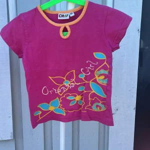 Rosa blommig topp från Ciraf - Söt rosa topp från Ciraf med korta ärmar och orange kant vid halsen. Framsidan har färgglada blommor i turkos och gult samt texten 'Oriental Girl'. Nyckelhålsdetalj vid halsringningen. Perfekt för dig som gillar färg och lekfull stil.