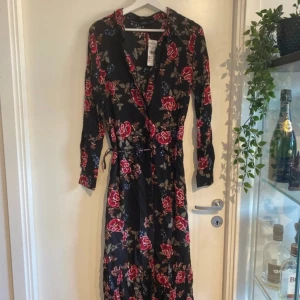 Svart blommig omlottklänning från KappAhl - Svart långklänning från KappAhl med röda och rosa blommor, omlottmodell och knytband i midjan. Klänningen har lång ärm och v-ringad hals. Perfekt för dig som gillar romantiska mönster och vill ha en feminin look. Nypris 400kr