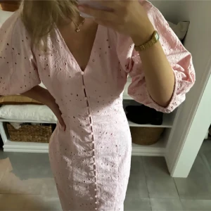 Ljusrosa klänning med puffärmar och broderade detaljer - Superfin ljusrosa klänning med v-ringning, puffiga trekvartsärmar och broderade hålmönster. Klänningen har knappar framtill och markerad midja för en feminin siluett. Perfekt för sommarens alla tillfällen!