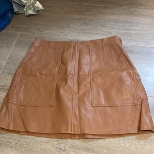 Brun skinnkjol från H&M - Säljer en snygg brun skinnkjol från H&M i storlek 42. Kjolen har två stora fickor framtill och en rak passform. Perfekt att styla med en enkel topp eller stickad tröja.