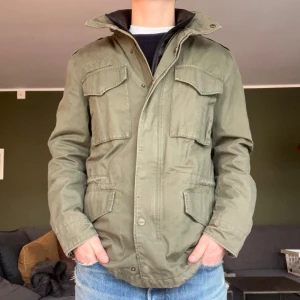 Grön Fieldjacka från Dobber - Säljer en grön Fieldjacka från Dobber med dragkedja och knappar. Jackan har flera praktiska fickor och en hög krage. Perfekt för våren som kommer. Sitter mer som en Medium.