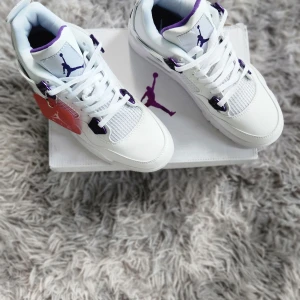 Nike Air Jordan 4 Retro 'Court Purple' - Säljer ett par fräscha vita Nike Air Jordan 4 Retro med lila detaljer och klassisk Jumpman-logga. Skorna har snörning, meshpaneler och en platt sula. Perfekta för dig som vill sticka ut med stilrena sneakers.