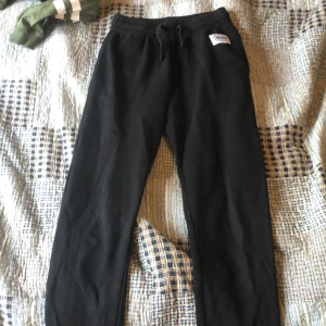 Svarta mjukisbyxor med dragsko - Svarta mjukisbyxor med elastisk midja och dragsko. Byxorna har en enkel design och en liten vit patch vid fickan. Perfekta för en avslappnad stil.