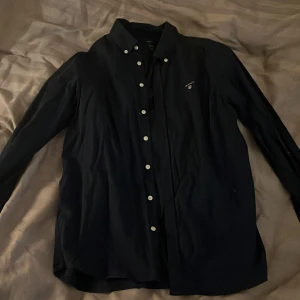 Mörkblå skjorta från GANT - Säljer en klassisk mörkblå skjorta från GANT med vita knappar och broderad logga på bröstet. Skjortan har lång ärm och är perfekt för dig som gillar stilrena plagg med snygga detaljer.