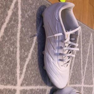 Vita fotbollsskor från Adidas - Säljer ett par vita fotbollsskor från Adidas med snörning och rund tå. Skorna har gröna dobbar och är tillverkade i syntetmaterial. Perfekta för dig som vill ha stilrena och funktionella skor på planen. Medföljer extra dobbar och verktyg. Storlek 41 1/3