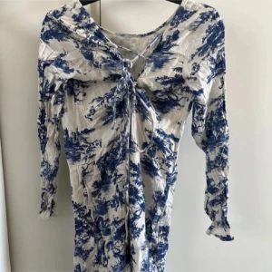 Vit och blå långärmad klänning med knyt - Säljer en vit klänning med blått mönster och långa ärmar. Klänningen har v-ringning och knyt framtill, vilket ger en snygg detalj. Strl S