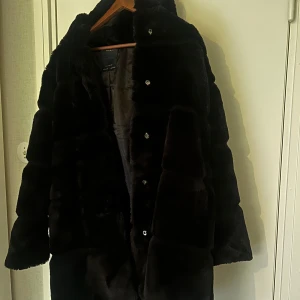 Svart pälsjacka med knappar - Säljer en svart pälsjacka med knäppning framtill. Jackan har lång ärm och en klassisk, lite oversized passform. Perfekt för kalla dagar när du vill vara både varm och snygg.
