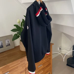 Svart hoodie med dragkedja från Moncler - Svart hoodie med dragkedja från Moncler. Tröjan har huva, fickor framtill och ribbade muddar med röd och vit rand. Moncler-logga på ärmen och sportig känsla med detaljer i rött och vitt.