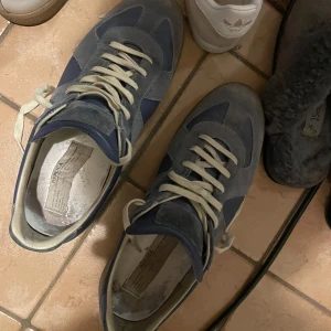 Blå och grå sneakers från Maison Margiela - Säljer ett par klassiska sneakers från Maison Margiela i blått och grått med vita snören och gummisula. Skorna har en mix av mocka och skinn, rund tå och platt sula. Perfekta för dig som gillar stilrena och tidlösa sneakers.Färgen pewter, dem enda äkta på marknaden. Har kvitto. Öppen till byten, men måste då gynna mig. Detta är en Intressekoll, kommer inte ge bort dem för låg pris