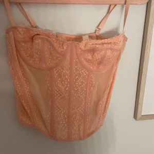 Persikofärgad spetskorsett - Säljer en persikofärgad korsett med vackra spetsdetaljer och justerbara axelband. Toppen har en feminin design med genomskinliga partier och dekorativa sömmar framtill. Perfekt för att lyfta vilken outfit som helst!