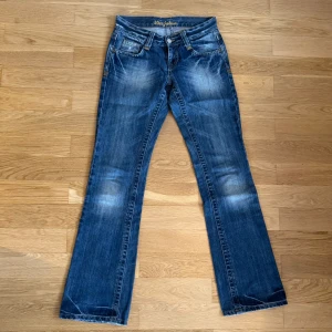 Blå bootcut jeans från Miss Julien - Snygga blå bootcut jeans från Miss Julien med låg midja. Slitningar framtill och en broderad palm på baksidan av linningen. Ca 36 cm rakt över midjan och 80 cm i innerbenslängd. Inte så stretchiga med mer ”ordentligt” jeanstyg.