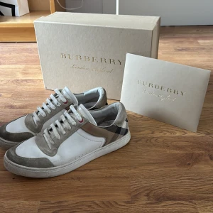 Burberry skor - Säljer ett par snygga Burberry sneakers i storlek 42,5., skick 7/10 - Vita och beige med klassiskt rutigt mönster på hälen. Skorna har snörning, rund tå och platt sula. Perfekta för dig som gillar stilrena och exklusiva sneakers med en touch av lyx. Hör av er vid frågor 😊