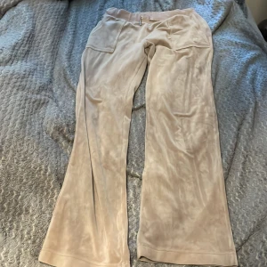Beiga velourbyxor från Juicy Couture - Säljer ett par beiga velourbyxor från Juicy Couture i storlek M. Byxorna har bred resårmidja med snörning, fickor fram och bak med knappdetaljer och en mjuk, glansig finish. Perfekta för en avslappnad och trendig look.