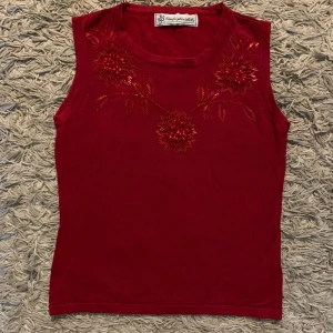 Röd ärmlös topp med broderade blommor - Snygg röd ärmlös topp med broderade blommor och pärlor framtill. Perfekt för att piffa upp din outfit med lite extra detalj. Mjukt material och rund halsringning.