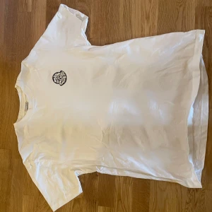 Vit t-shirt från Moncler med grafiskt tryck - Säljer en vit t-shirt från Moncler med ett stort färgglatt grafiskt tryck och Moncler-logga på ryggen. Framtill finns en broderad logga på bröstet och en liten patch på ärmen. Klassisk passform och rund hals.