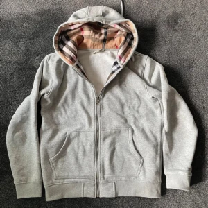Grå hoodie från Burberry med klassiskt rutigt foder - Säljer en grå hoodie från Burberry med dragkedja och kängurufickor. Huvan är fodrad med Burberrys ikoniska rutiga tyg och tröjan har diskret broderad logga på bröstet. Perfekt för en stilren och avslappnad look. Fin inför sommarn, inte 100% om den är äkta samt så strular dragkedjan mycket, annaes fint skick!!