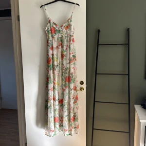 Blommig långklänning med smala axelband - Säljer en romantisk långklänning med smala, justerbara axelband och v-ringning. Klänningen har ett vackert blommigt mönster i grönt, rött, gult och orange på vit botten. Lätt och luftig modell, perfekt för varma dagar.