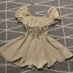 Beige klänning med puffärmar och broderad midja - Supersöt beige klänning med puffiga kortärmar och broderad midja med blommönster och snörning framtill. Klänningen har en lätt utställd kjol och romantisk känsla, perfekt för sommarens alla tillfällen.