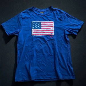 Blå t-shirt med amerikansk flagga från Polo Ralph Lauren - Säljer en blå t-shirt från Polo Ralph Lauren med en broderad amerikansk flagga på bröstet och RL-broderi. Klassisk passform och mjukt bomullsmaterial. Perfekt för dig som gillar stilrena och ikoniska plagg. Som ni ser har den ett litet hål vid flaggan därför det billiga priset skick 7,5/10