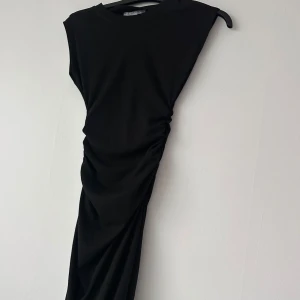 Svart långklänning med rynkade detaljer - Säljer en svart långklänning med snygga rynkade detaljer längs sidan. Klänningen har en enkel och stilren design med ärmlös modell och rund halsringning. Perfekt för dig som gillar minimalistisk stil.