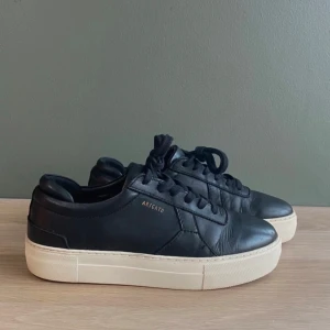 Axel Arigato sneakers  - Snygga svarta sneakers från Axel Arigato med vit platt sula och diskret guldfärgad logga på sidan. Skorna har snörning och är tillverkade i skinn, vilket ger en stilren och clean look. Perfekta för dig som gillar minimalistisk stil. Bra skick, inga defekter💕