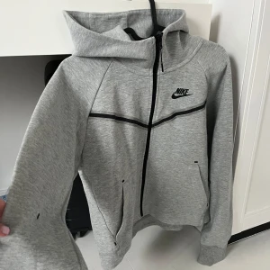 Nike Tech Fleece women  - Säljer en grå Tech Fleece från Nike med svart logga på bröstet och svarta detaljer längs dragkedjan. Den har luva, två fickor framtill och långa ärmar. Perfekt för en sportig och avslappnad stil. Tillverkad i tjockt lyxigt material. Använd Max en gång så den är som ny! Köpte den för 1,500kr 💓