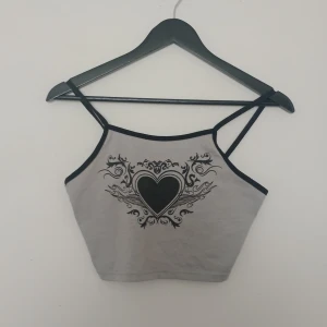 Grå croppad linnetopp med hjärtmotiv - Säljer en grå croppad linnetopp från Romwe med svarta tunna axelband och ett stort svart hjärtmotiv med tribal-inspirerade detaljer framtill. Perfekt för en alternativ stil.