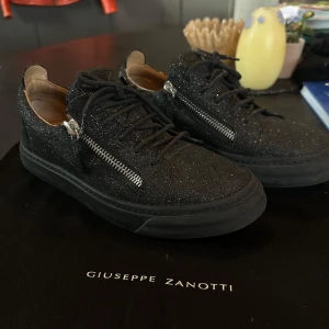 Svarta glittriga sneakers från Giuseppe Zanotti - Säljer ett par svarta sneakers från Giuseppe Zanotti med glittrig yta och silverfärgade dragkedjor på sidorna. Skorna har rund tå, platt sula och klassisk snörning. Skorna är tillverkade i Italien och är perfekta för dig som vill sticka ut med stilrena detaljer. Är i väldigt bra skick endast lite slitna på undersidan men inget hål eller sådant. Skulle säga att skorna är ganska stora i storleken, jag som har 38/39 kan använda dem men de är stora. Skulle säga att de är bättre med strl. 39,5-40,5✨🖤