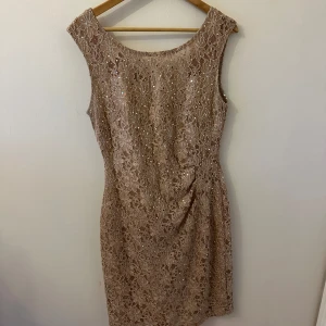 Champagnefärgad spetsklänning - Säljer en elegant champagnefärgad klänning med vackert blommigt spetsmönster och glittriga detaljer. Klänningen är ärmlös och har en figurnära passform med rundad halsringning. Perfekt för speciella tillfällen!