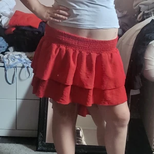 Röd volangkjol med resårmidja - Säljer en kort röd kjol med två volanglager och bred resår i midjan. Kjolen har en luftig och flowy känsla, perfekt för dig som vill ha något färgstarkt och bekvämt. Passar snyggt till både enkla toppar och stickade tröjor.