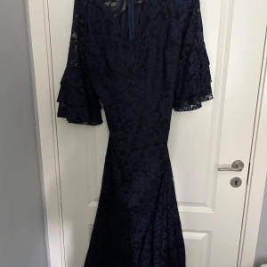 Marinblå långklänning med spets och volangärmar - Elegant marinblå långklänning med vackert spetsmönster och vida volangärmar. Klänningen har en figurnära passform upptill och faller snyggt längs kroppen. Perfekt för festliga tillfällen där du vill sticka ut med stil.