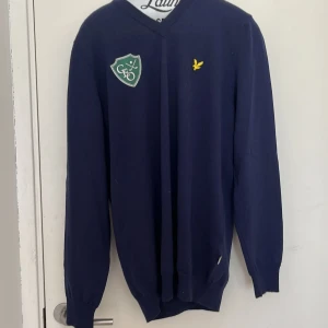 Mörkblå v-ringad tröja från Lyle & Scott Golf - Mörkblå v-ringad tröja från Lyle & Scott Golf med lång ärm. Tröjan har en broderad gul logotyp på bröstet och en grön patch på vänster sida. Perfekt för golf eller vardag.