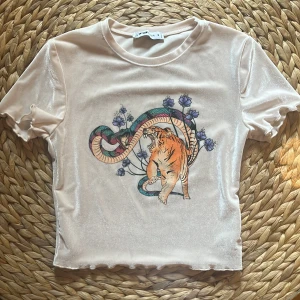  kortärmad topp med tiger- och ormmotiv från FB SISTER - Säljer en glittrig ljusbeige topp från FB SISTER med korta ärmar. Framsidan har ett stort tryck med en tiger, en orm och lila blommor. Tyget har en skimrande finish och toppen är croppad med lätt vågig kant.