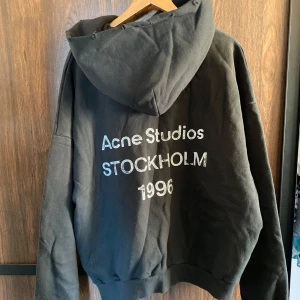 Svart hoodie från Acne Studios - Svart hoodie från Acne Studios med tryck på ryggen: 'Acne Studios STOCKHOLM 1996'. Klassisk modell med huva och ribbade muddar. Perfekt för en avslappnad och stilren look. Är storlek S men skulle säga att den passar M bättre. Hör av dig vid minsta fråga!