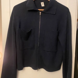 Mörkblå stickad jacka med dragkedja från Å - Mörkblå stickad jacka från Å med dragkedja framtill och två stora fickor på bröstet. Klassisk krage och långärmad modell, perfekt för lager-på-lager. Enkel och stilren design som passar till mycket.