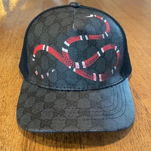 Svart keps från Gucci med ormtryck - Snygg svart keps från Gucci med diskret GG-mönster och en röd-vit orm på framsidan. Justerbar rem baktill med blå och röd rand. Kepsen är tillverkad i Italien och har mesh på baksidan för extra ventilation.