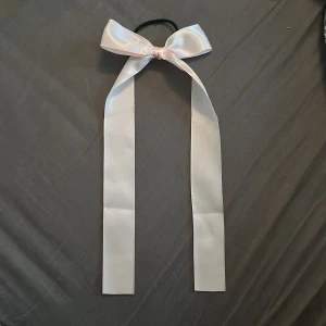 Snodd med rosett - Svart snodd med en lång, glansig rosett i satin. Perfekt accessoar för att piffa upp frisyren med en söt och stilren detalj. Rosetten sitter på ett elastiskt band som gör det enkelt att använda.
