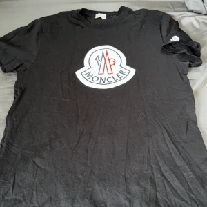 Svart t-shirt från Moncler med stor logga - Säljer en svart t-shirt från Moncler med en stor broderad logga i vitt, rött och blått på bröstet. Klassisk passform och korta ärmar. Perfekt för dig som gillar stilrena märkesplagg. Betala genom Swish 