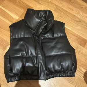 Svart dunväst från Zara - Snygg svart dunväst från Zara med glansig finish och hög krage. Västen har dragkedja och tryckknappar framtill samt elastisk nederkant för en modern look. Perfekt att styla över en hoodie eller tröja.