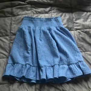 Blå volangkjol från Stories - Supersöt blå kjol från Stories i storlek XS. Kjolen har en smockad midja och volang nertill som ger en fin och lekfull look. Perfekt för varma dagar och enkel att matcha med olika toppar.